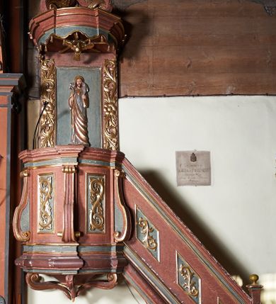 Zdjęcie nr 1: Ambona zawieszona na północnej ścianie prezbiterium. Kosz na rzucie owalu, z wydzielonym profilowanym cokołem i gzymsem, opięty wolutami umieszczonymi na tle pilastrów; wsparty na nadwieszonej podstawie o formie odwróconego spłaszczonego stożka, ujętego pięcioma wolutowo wygiętymi kabłąkami. Zaplecek prostokątny, łączący się z owalnym baldachimem zwieńczonym krzyżem ustawionym na czterech kabłąkach. Baldachim od dołu dekorowany lambrekinem. Od strony wschodniej schody z balustradą zdobioną romboidalnymi płycinami z aplikowanym ornamentem roślinnym. Płyciny kosza oraz boki zaplecka dekorowane wicią akantu. Pośrodku zaplecka płaskorzeźbiona figura Chrystusa Dobrego Pasterza, w podniebiu rzeźbiona Gołębica Ducha Świętego otoczona promieniami. Całość struktury ambony polichromowana na kolor ciemnobrązowy oraz ciemnozielony w partii płycin, detal i profilowania pozłacane.