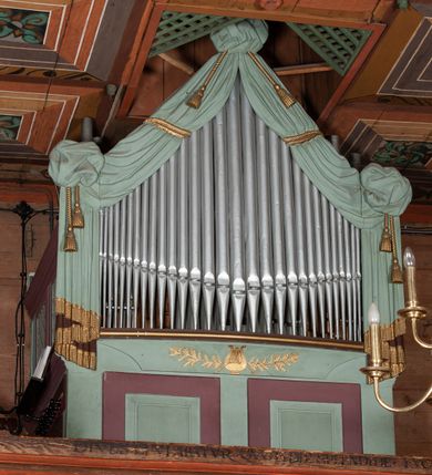 Zdjęcie nr 3: Prospekt organowy prostopadłościenny, z wydzielonym cokołem, jednoosiowy, jednokondygnacyjny. Część cokołowa podzielona prostokątnymi płycinami. Cześć górna z piszczałkami od czoła lekko wypukła, zwieńczona trójkątnie, ujęta rzeźbioną kotarą zakończoną frędzlami, podwiązaną pośrodku i po bokach złotymi sznurami. Poniżej, pośrodku aplikowana dekoracja z motywem liry przeplecionej gałązką oliwną. Na bokach prospektu ramy wypełnione ukośną kratką. Całość struktury polichromowana na jasnozielony kolor, profilowania ciemnobrązowe, detal architektoniczny pozłacany