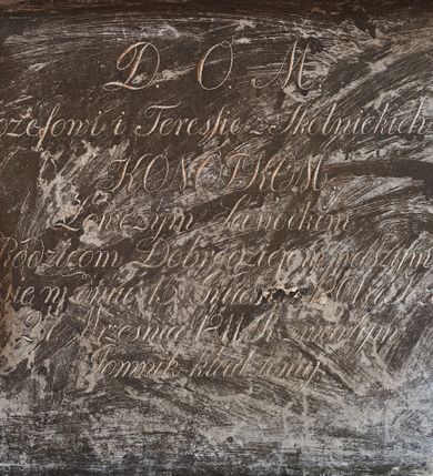 Zdjęcie nr 6: Wmurowany w ścianę kościoła nagrobek, na wysokim postumencie, z tablicą w kształcie leżącego prostokąta z czterema metalowymi ćwiekami w kształcie rozet, w szerokiej ramie z stiukowymi herbami Nowina i Bogoria na bokach i profilowanym gzymsem z czterema stiukowymi rozetami. Ponad gzymsem ukazana z prawego profilu klęcząca i wsparta na urnie kobieta, ubrana w togę i płaszcz zarzucony na głowę, plecy i częściowo zakrywający urnę, na której kobieta wspiera ręce i głowę. Twarz pełna, o delikatnych rysach i zamkniętych oczach. Włosy sięgające ramion, o gęstych, kręconych puklach. Urna w typie amfory greckiej zdobiona dołem szerokimi liśćmi i pięciopłatkowymi kwiatami, górą fryzem z liśćmi akantu. Na tablicy inskrypcja: &quot;D(eo) O(ptimo) M(aximo) / Józefowi i Teressie Skotnickich / KONOPKOM. / Łowczym Sanockim / Rodzicom Dobrodziejom Naszym. / Teressie w dniu 15. Grudnia 1801. a Józefowi / d(nia) 27. Września 1811. R(oku) zmarłym / Pomnik kładziemy&quot;. 