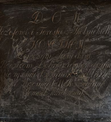 Zdjęcie nr 5: Wmurowany w ścianę kościoła nagrobek, na wysokim postumencie, z tablicą w kształcie leżącego prostokąta z czterema metalowymi ćwiekami w kształcie rozet, w szerokiej ramie z stiukowymi herbami Nowina i Bogoria na bokach i profilowanym gzymsem z czterema stiukowymi rozetami. Ponad gzymsem ukazana z prawego profilu klęcząca i wsparta na urnie kobieta, ubrana w togę i płaszcz zarzucony na głowę, plecy i częściowo zakrywający urnę, na której kobieta wspiera ręce i głowę. Twarz pełna, o delikatnych rysach i zamkniętych oczach. Włosy sięgające ramion, o gęstych, kręconych puklach. Urna w typie amfory greckiej zdobiona dołem szerokimi liśćmi i pięciopłatkowymi kwiatami, górą fryzem z liśćmi akantu. Na tablicy inskrypcja: "D(eo) O(ptimo) M(aximo) / Józefowi i Teressie Skotnickich / KONOPKOM. / Łowczym Sanockim / Rodzicom Dobrodziejom Naszym. / Teressie w dniu 15. Grudnia 1801. a Józefowi / d(nia) 27. Września 1811. R(oku) zmarłym / Pomnik kładziemy". 