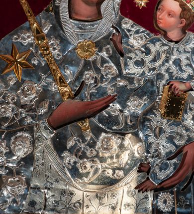 Zdjęcie nr 2: Metalowa sukienka na obraz Matki Boskiej z Dzieciątkiem w ołtarzu głównym, w kształcie obrysu sylwetek Marii i Dzieciątka, z otworami na twarze, dłonie i stopy Jezusa, zdobiona dekoracją roślinną i sześcioramienną złotą gwiazdą umieszczoną na prawym ramieniu Marii. W prawym dolnym narożu fragment sukienki o odmiennej dekoracji (być może pozostałość ze starszej sukienki). Korony w typie zamkniętym, zwieńczone krzyżykami, złocone, kameryzowane. Wokół głowy Matki Boskiej wieniec z gwiazd, w ręku złocone berło. Tło z nowego czerwonego aksamitu.
