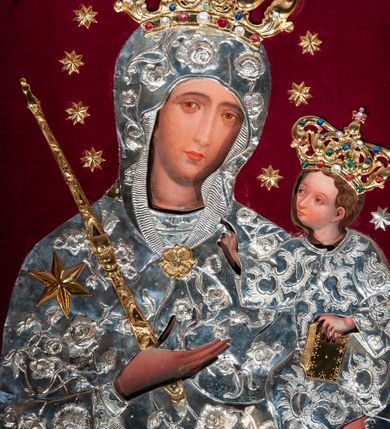 Zdjęcie nr 1: Metalowa sukienka na obraz Matki Boskiej z Dzieciątkiem w ołtarzu głównym, w kształcie obrysu sylwetek Marii i Dzieciątka, z otworami na twarze, dłonie i stopy Jezusa, zdobiona dekoracją roślinną i sześcioramienną złotą gwiazdą umieszczoną na prawym ramieniu Marii. W prawym dolnym narożu fragment sukienki o odmiennej dekoracji (być może pozostałość ze starszej sukienki). Korony w typie zamkniętym, zwieńczone krzyżykami, złocone, kameryzowane. Wokół głowy Matki Boskiej wieniec z gwiazd, w ręku złocone berło. Tło z nowego czerwonego aksamitu.