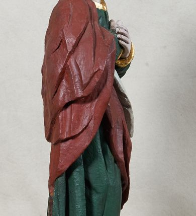 Zdjęcie nr 3: Rzeźba św. Jana Ewangelisty, pełnoplastyczna, z tyłu opracowana, ustawiona na niskim, prostopadłościennym postumencie.  Święty ukazany frontalnie, w nieznacznym kontrapoście, z prawą nogą ugiętą w kolanie, z dłońmi skrzyżowanymi na piersi, z głową przechyloną na prawy bark. Ubrany w długą zieloną suknię z długimi rękawami, ze złotą lamówką na brzegach, oraz czerwony płaszcz z białą podszewką zarzucony na prawe ramię, okrywający plecy i przełożony przez przód figury. Twarz szeroka; lekko rozchylone, wąskie usta; duże i migdałowate oczy. Włosy sięgające do ramion, delikatnie falowane. Szaty silnie drapowane.