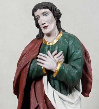 Zdjęcie nr 1: Rzeźba św. Jana Ewangelisty, pełnoplastyczna, z tyłu opracowana, ustawiona na niskim, prostopadłościennym postumencie.  Święty ukazany frontalnie, w nieznacznym kontrapoście, z prawą nogą ugiętą w kolanie, z dłońmi skrzyżowanymi na piersi, z głową przechyloną na prawy bark. Ubrany w długą zieloną suknię z długimi rękawami, ze złotą lamówką na brzegach, oraz czerwony płaszcz z białą podszewką zarzucony na prawe ramię, okrywający plecy i przełożony przez przód figury. Twarz szeroka; lekko rozchylone, wąskie usta; duże i migdałowate oczy. Włosy sięgające do ramion, delikatnie falowane. Szaty silnie drapowane.