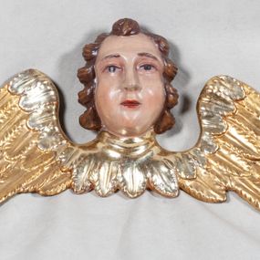 Zdjęcie nr 1: Główka aniołka ukazana frontalnie z szeroko rozpostartymi skrzydłami. Główka o owalnej twarzy, z pucułowatymi policzkami, małymi i wąskimi ustami, długim i szerokim nosem oraz wzrokiem skierowanym na wprost. Głowę okalają włosy z lokami zaczesanymi do tyłu i dużym loczkiem pośrodku nad czołem. Karnacja twarzy jasna, skrzydła złocone i srebrzone.