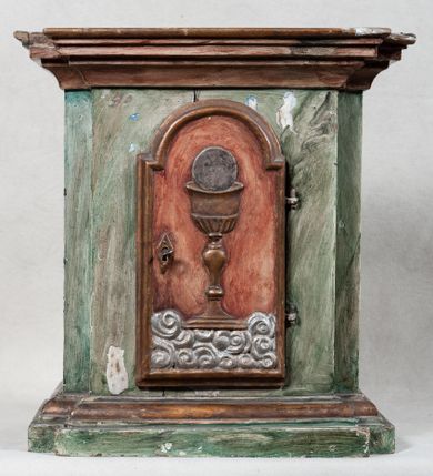 Zdjęcie nr 1: Tabernakulum w formie prostopadłościennej szafki o ściętych od przodu narożach, na niskim, profilowanym cokole, zamknięte od góry profilowanym gzymsem.  Drzwiczki w kształcie stojącego prostokąta zamkniętego łukiem nadwieszonym, osadzone na zawiasach, zamykane na zamek, ozdobione płaskorzeźbionym kielichem z hostią, stojącym na obłokach. Tło drzwiczek czerwone. Szyld zamka w kształcie wydłużonego rombu. Struktura marmoryzowana na kolor zielony, profilowania oraz detale złocone i srebrzone.