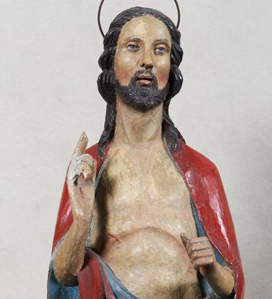 Zdjęcie nr 1: Rzeźba Chrystusa Zmartwychwstałego, pełnoplastyczna, z tyłu opracowana, ustawiona na niskim, okrągłym cokole. Chrystus ukazany w całej postaci, frontalnie, z prawą ręką uniesioną w geście błogosławieństwa, w lewej trzyma chorągiew. Ubrany w czerwony płaszcz z niebieską podszewką, spływający z lewego ramienia, przez przód postaci, na prawe przedramię, tworząc znaczną fałdę misową. Korpus ciała nagi. Twarz podłużna, z lekko rozchylonymi ustami, okolona krótko przystrzyżoną brodą i ciemnobrązowymi włosami opadającymi na plecy. Wokół głowy złoty, kolisty nimb z metalowego pręta. Czerwona chorągiew z tkaniny zawieszona na długim, złotym i drewnianym drzewcu zakończonym krzyżykiem.
