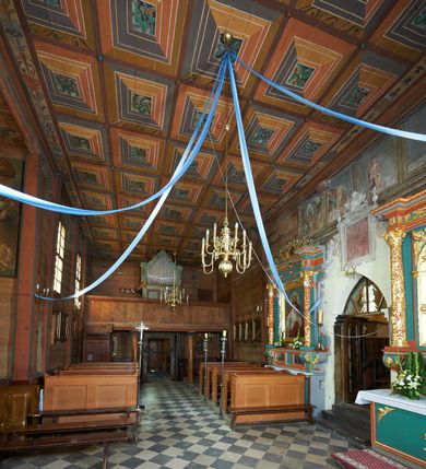 Zdjęcie nr 27: Prezbiterium. Strop pokryty dekoracją malarską imitującą kasetony wypełnione rozetami oraz umieszczonymi pomiędzy nimi krzyżami wypełnionymi liśćmi na ramionach i rozetami na skrzyżowaniu. Na ścianach bocznych prezbiterium, w arkadach podtrzymywanych przez kolumny popiersia apostołów oraz popiersia mężczyzn w strojach szlacheckich ukazane na tle zielonych obić (genealogia Chrystusa). Pod nimi fryz z łacińskimi napisami od dołu zdobiony girlandami z uskrzydlonymi główkami anielskimi oraz puklowanymi wazami. Na ścianach również herby: m.in. Łabędź, Jastrzębiec.  Nawa główna. Strop pokryty dekoracją malarską imitującą kasetony z rozetami. Na ścianach bocznych nawy głównej: na ścianie północnej cykl pasyjny w kwadratowych obramieniach z przedstawieniami: Modlitwa w Ogrojcu, Biczowanie Chrystusa, Chrystus Umęczony, Ecce Homo, Upadek pod krzyżem oraz słabo czytelny cykl maryjny z przedstawieniami: Zwiastowanie, Boże Narodzenie, Hołd Trzech Króli oraz Ucieczka do Egiptu i Rzeź niewiniątek, Jezus nauczający w świątyni. Na ścianie południowej nawy w arkadach umieszczono przedstawienia św. Wojciecha z wiosłem i św. Stanisława z Piotrowinem. Na balustradzie chóru muzycznego zachowały się częściowo trzy sceny ilustrujące przypowieść o bogaczu i Łazarzu oraz data: 1562. Kolorystyka pastelowa z przewagą zieleni, beżu i czerwieni z mocno zaznaczonymi konturami.