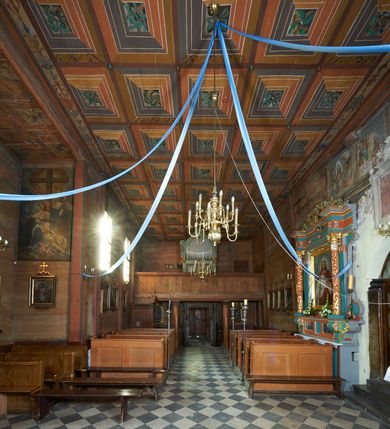 Zdjęcie nr 29: Prezbiterium. Strop pokryty dekoracją malarską imitującą kasetony wypełnione rozetami oraz umieszczonymi pomiędzy nimi krzyżami wypełnionymi liśćmi na ramionach i rozetami na skrzyżowaniu. Na ścianach bocznych prezbiterium, w arkadach podtrzymywanych przez kolumny popiersia apostołów oraz popiersia mężczyzn w strojach szlacheckich ukazane na tle zielonych obić (genealogia Chrystusa). Pod nimi fryz z łacińskimi napisami od dołu zdobiony girlandami z uskrzydlonymi główkami anielskimi oraz puklowanymi wazami. Na ścianach również herby: m.in. Łabędź, Jastrzębiec.  Nawa główna. Strop pokryty dekoracją malarską imitującą kasetony z rozetami. Na ścianach bocznych nawy głównej: na ścianie północnej cykl pasyjny w kwadratowych obramieniach z przedstawieniami: Modlitwa w Ogrojcu, Biczowanie Chrystusa, Chrystus Umęczony, Ecce Homo, Upadek pod krzyżem oraz słabo czytelny cykl maryjny z przedstawieniami: Zwiastowanie, Boże Narodzenie, Hołd Trzech Króli oraz Ucieczka do Egiptu i Rzeź niewiniątek, Jezus nauczający w świątyni. Na ścianie południowej nawy w arkadach umieszczono przedstawienia św. Wojciecha z wiosłem i św. Stanisława z Piotrowinem. Na balustradzie chóru muzycznego zachowały się częściowo trzy sceny ilustrujące przypowieść o bogaczu i Łazarzu oraz data: 1562. Kolorystyka pastelowa z przewagą zieleni, beżu i czerwieni z mocno zaznaczonymi konturami.