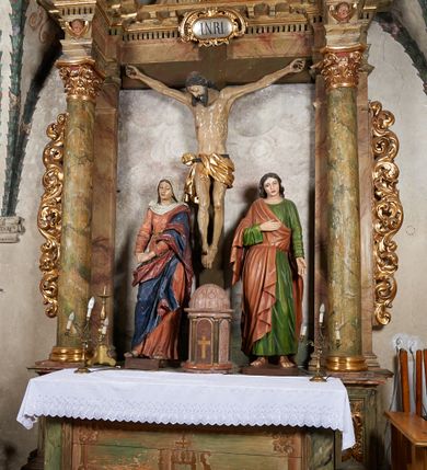 Zdjęcie nr 13: W centrum krzyż z figurą Chrystusa umarłego. Postać w lekkim zwisie o szeroko rozłożonych ramionach, głowa opada na prawe ramie. Twarz szczupła, wyraźnie rzeźbiona, okolona krótką brodą; oczy zamknięte, długi nos, usta delikatnie rozchylone. Włosy w postaci delikatnych loków opadają na ramię i plecy. Ciało szczupłe, ascetyczne, o plastycznie kształtowanej anatomii, z zaznaczonymi śladami męki. Nogi ugięte w kolanach, stopy przebite jednym gwoździem, założone prawa na lewą. Perizonium przewiązane podwójnym sznurem, odsłaniające prawe biodro, ze zwisem na prawym boku. Po prawej stronie krzyża figura Matki Boskiej, zwrócona en face, w kontrapoście; dłonie splecione, wyciągnięte przed siebie. Twarz okrągła o wyraźnych liniach łuków brwiowych i nosa, półprzymkniętych oczach. Ubrana w różową suknię przewiązaną w talii oraz granatowy płaszcz podbity czerwoną podszewką, zarzucony diagonalnie na lewe ramię i opadający na prawą nogę. Na głowie biały welon. Po lewej stronie krzyża figura św. Jana, zwrócona en face, w kontrapoście, o esowato wygiętej sylwetce; prawą rękę trzyma na piersi, lewa pozostaje opuszczona wzdłuż ciała. Twarz pełna, młodzieńcza, podkreślone linie łuków brwiowych i nosa. Włosy długie, falowane, opadają na ramiona. Ubrany w zieloną tunikę oraz czerwony płaszcz przerzucony diagonalnie przez prawy bok, przewiązane w talii. Polichromia naturalistyczna, perizonium złocone.Grupa na tle malowanego miasta oraz skłębionego szaroróżowego nieba.