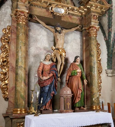 Zdjęcie nr 2: W centrum krzyż z figurą Chrystusa umarłego. Postać w lekkim zwisie o szeroko rozłożonych ramionach, głowa opada na prawe ramie. Twarz szczupła, wyraźnie rzeźbiona, okolona krótką brodą; oczy zamknięte, długi nos, usta delikatnie rozchylone. Włosy w postaci delikatnych loków opadają na ramię i plecy. Ciało szczupłe, ascetyczne, o plastycznie kształtowanej anatomii, z zaznaczonymi śladami męki. Nogi ugięte w kolanach, stopy przebite jednym gwoździem, założone prawa na lewą. Perizonium przewiązane podwójnym sznurem, odsłaniające prawe biodro, ze zwisem na prawym boku. Po prawej stronie krzyża figura Matki Boskiej, zwrócona en face, w kontrapoście; dłonie splecione, wyciągnięte przed siebie. Twarz okrągła o wyraźnych liniach łuków brwiowych i nosa, półprzymkniętych oczach. Ubrana w różową suknię przewiązaną w talii oraz granatowy płaszcz podbity czerwoną podszewką, zarzucony diagonalnie na lewe ramię i opadający na prawą nogę. Na głowie biały welon. Po lewej stronie krzyża figura św. Jana, zwrócona en face, w kontrapoście, o esowato wygiętej sylwetce; prawą rękę trzyma na piersi, lewa pozostaje opuszczona wzdłuż ciała. Twarz pełna, młodzieńcza, podkreślone linie łuków brwiowych i nosa. Włosy długie, falowane, opadają na ramiona. Ubrany w zieloną tunikę oraz czerwony płaszcz przerzucony diagonalnie przez prawy bok, przewiązane w talii. Polichromia naturalistyczna, perizonium złocone.Grupa na tle malowanego miasta oraz skłębionego szaroróżowego nieba.