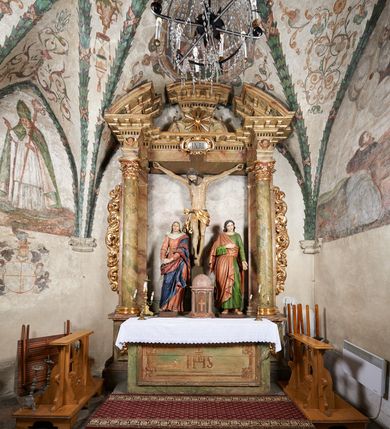 Zdjęcie nr 11: W centrum krzyż z figurą Chrystusa umarłego. Postać w lekkim zwisie o szeroko rozłożonych ramionach, głowa opada na prawe ramie. Twarz szczupła, wyraźnie rzeźbiona, okolona krótką brodą; oczy zamknięte, długi nos, usta delikatnie rozchylone. Włosy w postaci delikatnych loków opadają na ramię i plecy. Ciało szczupłe, ascetyczne, o plastycznie kształtowanej anatomii, z zaznaczonymi śladami męki. Nogi ugięte w kolanach, stopy przebite jednym gwoździem, założone prawa na lewą. Perizonium przewiązane podwójnym sznurem, odsłaniające prawe biodro, ze zwisem na prawym boku. Po prawej stronie krzyża figura Matki Boskiej, zwrócona en face, w kontrapoście; dłonie splecione, wyciągnięte przed siebie. Twarz okrągła o wyraźnych liniach łuków brwiowych i nosa, półprzymkniętych oczach. Ubrana w różową suknię przewiązaną w talii oraz granatowy płaszcz podbity czerwoną podszewką, zarzucony diagonalnie na lewe ramię i opadający na prawą nogę. Na głowie biały welon. Po lewej stronie krzyża figura św. Jana, zwrócona en face, w kontrapoście, o esowato wygiętej sylwetce; prawą rękę trzyma na piersi, lewa pozostaje opuszczona wzdłuż ciała. Twarz pełna, młodzieńcza, podkreślone linie łuków brwiowych i nosa. Włosy długie, falowane, opadają na ramiona. Ubrany w zieloną tunikę oraz czerwony płaszcz przerzucony diagonalnie przez prawy bok, przewiązane w talii. Polichromia naturalistyczna, perizonium złocone.Grupa na tle malowanego miasta oraz skłębionego szaroróżowego nieba.