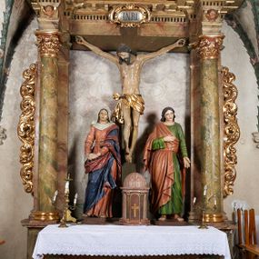 Zdjęcie nr 1: W centrum krzyż z figurą Chrystusa umarłego. Postać w lekkim zwisie o szeroko rozłożonych ramionach, głowa opada na prawe ramie. Twarz szczupła, wyraźnie rzeźbiona, okolona krótką brodą; oczy zamknięte, długi nos, usta delikatnie rozchylone. Włosy w postaci delikatnych loków opadają na ramię i plecy. Ciało szczupłe, ascetyczne, o plastycznie kształtowanej anatomii, z zaznaczonymi śladami męki. Nogi ugięte w kolanach, stopy przebite jednym gwoździem, założone prawa na lewą. Perizonium przewiązane podwójnym sznurem, odsłaniające prawe biodro, ze zwisem na prawym boku. Po prawej stronie krzyża figura Matki Boskiej, zwrócona en face, w kontrapoście; dłonie splecione, wyciągnięte przed siebie. Twarz okrągła o wyraźnych liniach łuków brwiowych i nosa, półprzymkniętych oczach. Ubrana w różową suknię przewiązaną w talii oraz granatowy płaszcz podbity czerwoną podszewką, zarzucony diagonalnie na lewe ramię i opadający na prawą nogę. Na głowie biały welon. Po lewej stronie krzyża figura św. Jana, zwrócona en face, w kontrapoście, o esowato wygiętej sylwetce; prawą rękę trzyma na piersi, lewa pozostaje opuszczona wzdłuż ciała. Twarz pełna, młodzieńcza, podkreślone linie łuków brwiowych i nosa. Włosy długie, falowane, opadają na ramiona. Ubrany w zieloną tunikę oraz czerwony płaszcz przerzucony diagonalnie przez prawy bok, przewiązane w talii. Polichromia naturalistyczna, perizonium złocone.Grupa na tle malowanego miasta oraz skłębionego szaroróżowego nieba.