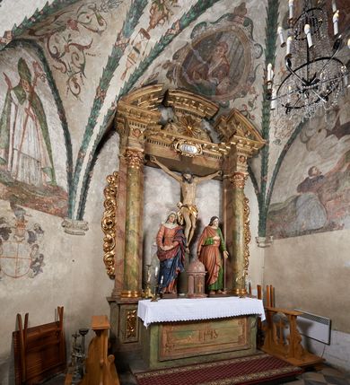 Zdjęcie nr 12: W centrum krzyż z figurą Chrystusa umarłego. Postać w lekkim zwisie o szeroko rozłożonych ramionach, głowa opada na prawe ramie. Twarz szczupła, wyraźnie rzeźbiona, okolona krótką brodą; oczy zamknięte, długi nos, usta delikatnie rozchylone. Włosy w postaci delikatnych loków opadają na ramię i plecy. Ciało szczupłe, ascetyczne, o plastycznie kształtowanej anatomii, z zaznaczonymi śladami męki. Nogi ugięte w kolanach, stopy przebite jednym gwoździem, założone prawa na lewą. Perizonium przewiązane podwójnym sznurem, odsłaniające prawe biodro, ze zwisem na prawym boku. Po prawej stronie krzyża figura Matki Boskiej, zwrócona en face, w kontrapoście; dłonie splecione, wyciągnięte przed siebie. Twarz okrągła o wyraźnych liniach łuków brwiowych i nosa, półprzymkniętych oczach. Ubrana w różową suknię przewiązaną w talii oraz granatowy płaszcz podbity czerwoną podszewką, zarzucony diagonalnie na lewe ramię i opadający na prawą nogę. Na głowie biały welon. Po lewej stronie krzyża figura św. Jana, zwrócona en face, w kontrapoście, o esowato wygiętej sylwetce; prawą rękę trzyma na piersi, lewa pozostaje opuszczona wzdłuż ciała. Twarz pełna, młodzieńcza, podkreślone linie łuków brwiowych i nosa. Włosy długie, falowane, opadają na ramiona. Ubrany w zieloną tunikę oraz czerwony płaszcz przerzucony diagonalnie przez prawy bok, przewiązane w talii. Polichromia naturalistyczna, perizonium złocone.Grupa na tle malowanego miasta oraz skłębionego szaroróżowego nieba.