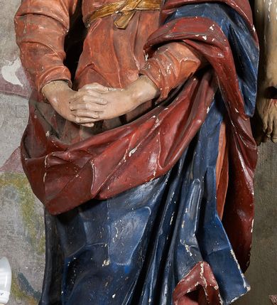 Zdjęcie nr 4: W centrum krzyż z figurą Chrystusa umarłego. Postać w lekkim zwisie o szeroko rozłożonych ramionach, głowa opada na prawe ramie. Twarz szczupła, wyraźnie rzeźbiona, okolona krótką brodą; oczy zamknięte, długi nos, usta delikatnie rozchylone. Włosy w postaci delikatnych loków opadają na ramię i plecy. Ciało szczupłe, ascetyczne, o plastycznie kształtowanej anatomii, z zaznaczonymi śladami męki. Nogi ugięte w kolanach, stopy przebite jednym gwoździem, założone prawa na lewą. Perizonium przewiązane podwójnym sznurem, odsłaniające prawe biodro, ze zwisem na prawym boku. Po prawej stronie krzyża figura Matki Boskiej, zwrócona en face, w kontrapoście; dłonie splecione, wyciągnięte przed siebie. Twarz okrągła o wyraźnych liniach łuków brwiowych i nosa, półprzymkniętych oczach. Ubrana w różową suknię przewiązaną w talii oraz granatowy płaszcz podbity czerwoną podszewką, zarzucony diagonalnie na lewe ramię i opadający na prawą nogę. Na głowie biały welon. Po lewej stronie krzyża figura św. Jana, zwrócona en face, w kontrapoście, o esowato wygiętej sylwetce; prawą rękę trzyma na piersi, lewa pozostaje opuszczona wzdłuż ciała. Twarz pełna, młodzieńcza, podkreślone linie łuków brwiowych i nosa. Włosy długie, falowane, opadają na ramiona. Ubrany w zieloną tunikę oraz czerwony płaszcz przerzucony diagonalnie przez prawy bok, przewiązane w talii. Polichromia naturalistyczna, perizonium złocone.Grupa na tle malowanego miasta oraz skłębionego szaroróżowego nieba.