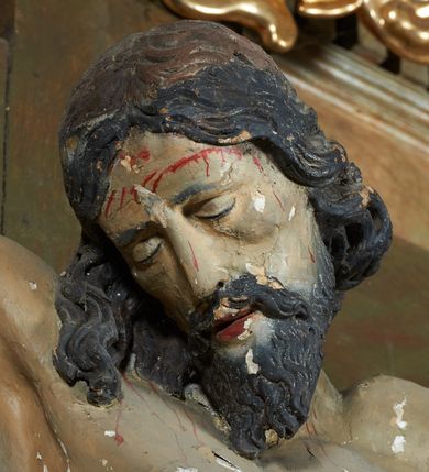 Zdjęcie nr 18: W centrum krzyż z figurą Chrystusa umarłego. Postać w lekkim zwisie o szeroko rozłożonych ramionach, głowa opada na prawe ramie. Twarz szczupła, wyraźnie rzeźbiona, okolona krótką brodą; oczy zamknięte, długi nos, usta delikatnie rozchylone. Włosy w postaci delikatnych loków opadają na ramię i plecy. Ciało szczupłe, ascetyczne, o plastycznie kształtowanej anatomii, z zaznaczonymi śladami męki. Nogi ugięte w kolanach, stopy przebite jednym gwoździem, założone prawa na lewą. Perizonium przewiązane podwójnym sznurem, odsłaniające prawe biodro, ze zwisem na prawym boku. Po prawej stronie krzyża figura Matki Boskiej, zwrócona en face, w kontrapoście; dłonie splecione, wyciągnięte przed siebie. Twarz okrągła o wyraźnych liniach łuków brwiowych i nosa, półprzymkniętych oczach. Ubrana w różową suknię przewiązaną w talii oraz granatowy płaszcz podbity czerwoną podszewką, zarzucony diagonalnie na lewe ramię i opadający na prawą nogę. Na głowie biały welon. Po lewej stronie krzyża figura św. Jana, zwrócona en face, w kontrapoście, o esowato wygiętej sylwetce; prawą rękę trzyma na piersi, lewa pozostaje opuszczona wzdłuż ciała. Twarz pełna, młodzieńcza, podkreślone linie łuków brwiowych i nosa. Włosy długie, falowane, opadają na ramiona. Ubrany w zieloną tunikę oraz czerwony płaszcz przerzucony diagonalnie przez prawy bok, przewiązane w talii. Polichromia naturalistyczna, perizonium złocone.Grupa na tle malowanego miasta oraz skłębionego szaroróżowego nieba.