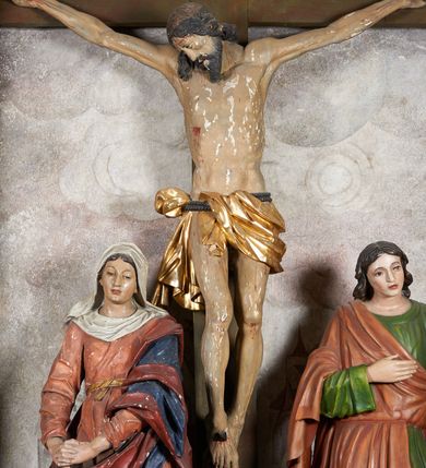 Zdjęcie nr 14: W centrum krzyż z figurą Chrystusa umarłego. Postać w lekkim zwisie o szeroko rozłożonych ramionach, głowa opada na prawe ramie. Twarz szczupła, wyraźnie rzeźbiona, okolona krótką brodą; oczy zamknięte, długi nos, usta delikatnie rozchylone. Włosy w postaci delikatnych loków opadają na ramię i plecy. Ciało szczupłe, ascetyczne, o plastycznie kształtowanej anatomii, z zaznaczonymi śladami męki. Nogi ugięte w kolanach, stopy przebite jednym gwoździem, założone prawa na lewą. Perizonium przewiązane podwójnym sznurem, odsłaniające prawe biodro, ze zwisem na prawym boku. Po prawej stronie krzyża figura Matki Boskiej, zwrócona en face, w kontrapoście; dłonie splecione, wyciągnięte przed siebie. Twarz okrągła o wyraźnych liniach łuków brwiowych i nosa, półprzymkniętych oczach. Ubrana w różową suknię przewiązaną w talii oraz granatowy płaszcz podbity czerwoną podszewką, zarzucony diagonalnie na lewe ramię i opadający na prawą nogę. Na głowie biały welon. Po lewej stronie krzyża figura św. Jana, zwrócona en face, w kontrapoście, o esowato wygiętej sylwetce; prawą rękę trzyma na piersi, lewa pozostaje opuszczona wzdłuż ciała. Twarz pełna, młodzieńcza, podkreślone linie łuków brwiowych i nosa. Włosy długie, falowane, opadają na ramiona. Ubrany w zieloną tunikę oraz czerwony płaszcz przerzucony diagonalnie przez prawy bok, przewiązane w talii. Polichromia naturalistyczna, perizonium złocone.Grupa na tle malowanego miasta oraz skłębionego szaroróżowego nieba.