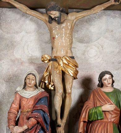 Zdjęcie nr 15: W centrum krzyż z figurą Chrystusa umarłego. Postać w lekkim zwisie o szeroko rozłożonych ramionach, głowa opada na prawe ramie. Twarz szczupła, wyraźnie rzeźbiona, okolona krótką brodą; oczy zamknięte, długi nos, usta delikatnie rozchylone. Włosy w postaci delikatnych loków opadają na ramię i plecy. Ciało szczupłe, ascetyczne, o plastycznie kształtowanej anatomii, z zaznaczonymi śladami męki. Nogi ugięte w kolanach, stopy przebite jednym gwoździem, założone prawa na lewą. Perizonium przewiązane podwójnym sznurem, odsłaniające prawe biodro, ze zwisem na prawym boku. Po prawej stronie krzyża figura Matki Boskiej, zwrócona en face, w kontrapoście; dłonie splecione, wyciągnięte przed siebie. Twarz okrągła o wyraźnych liniach łuków brwiowych i nosa, półprzymkniętych oczach. Ubrana w różową suknię przewiązaną w talii oraz granatowy płaszcz podbity czerwoną podszewką, zarzucony diagonalnie na lewe ramię i opadający na prawą nogę. Na głowie biały welon. Po lewej stronie krzyża figura św. Jana, zwrócona en face, w kontrapoście, o esowato wygiętej sylwetce; prawą rękę trzyma na piersi, lewa pozostaje opuszczona wzdłuż ciała. Twarz pełna, młodzieńcza, podkreślone linie łuków brwiowych i nosa. Włosy długie, falowane, opadają na ramiona. Ubrany w zieloną tunikę oraz czerwony płaszcz przerzucony diagonalnie przez prawy bok, przewiązane w talii. Polichromia naturalistyczna, perizonium złocone.Grupa na tle malowanego miasta oraz skłębionego szaroróżowego nieba.