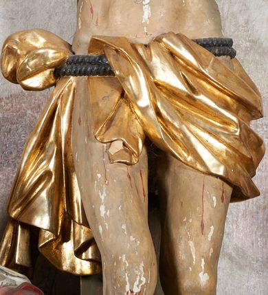 Zdjęcie nr 19: W centrum krzyż z figurą Chrystusa umarłego. Postać w lekkim zwisie o szeroko rozłożonych ramionach, głowa opada na prawe ramie. Twarz szczupła, wyraźnie rzeźbiona, okolona krótką brodą; oczy zamknięte, długi nos, usta delikatnie rozchylone. Włosy w postaci delikatnych loków opadają na ramię i plecy. Ciało szczupłe, ascetyczne, o plastycznie kształtowanej anatomii, z zaznaczonymi śladami męki. Nogi ugięte w kolanach, stopy przebite jednym gwoździem, założone prawa na lewą. Perizonium przewiązane podwójnym sznurem, odsłaniające prawe biodro, ze zwisem na prawym boku. Po prawej stronie krzyża figura Matki Boskiej, zwrócona en face, w kontrapoście; dłonie splecione, wyciągnięte przed siebie. Twarz okrągła o wyraźnych liniach łuków brwiowych i nosa, półprzymkniętych oczach. Ubrana w różową suknię przewiązaną w talii oraz granatowy płaszcz podbity czerwoną podszewką, zarzucony diagonalnie na lewe ramię i opadający na prawą nogę. Na głowie biały welon. Po lewej stronie krzyża figura św. Jana, zwrócona en face, w kontrapoście, o esowato wygiętej sylwetce; prawą rękę trzyma na piersi, lewa pozostaje opuszczona wzdłuż ciała. Twarz pełna, młodzieńcza, podkreślone linie łuków brwiowych i nosa. Włosy długie, falowane, opadają na ramiona. Ubrany w zieloną tunikę oraz czerwony płaszcz przerzucony diagonalnie przez prawy bok, przewiązane w talii. Polichromia naturalistyczna, perizonium złocone.Grupa na tle malowanego miasta oraz skłębionego szaroróżowego nieba.