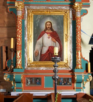 Zdjęcie nr 2: Obraz w kształcie stojącego prostokąta przedstawiający Chrystusa na tle jasnoszarych obłoków. Chrystus przedstawiony w ujęciu do kolan, stojący frontalnie, z prawą ręką opuszczoną, lekko odwiedzioną od tułowia i wyciągniętą do przodu. Lewą dłonią, spoczywającą na piersi, wskazuje na gorejące serce. Twarz Chrystusa pociągła, o jasnej karnacji, okolona krótkim zarostem i długimi włosami opadającymi na ramiona. Rysy twarzy wyraziste, oczy lekko przymknięte, nos szeroki, długi, usta niewielkie. Ubrany w białą suknię z długimi, szerokimi rękawami oraz czerwony płaszcz spięty na prawym ramieniu, z połami założonymi za szarfę wokół pasa. Z tyłu głowy świetlisty nimb krzyżowy.