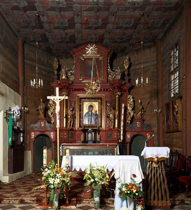 Zdjęcie nr 2:  Nastawa ołtarzowa architektoniczna, jednoosiowa, jednokondygnacyjna, na cokole, ze zwieńczeniem, z bramkami po bokach. Mensa drewniana z portatylem z marmuru dębnickiego, osadzona na murowanej, prostopadłościennej skrzyni. Antepedium w kształcie leżącego prostokąta, w profilowanej ramie z współczesnym obrazem Ostatnia Wieczerza, wzorowanym na fresku Leonarda da Vinci. Retabulum ujęte parą wklęsłych pilastrów oraz parą kolumn o kapitelach jońskich i gładkich trzonach, umieszczonych na ustawionych ukośnie cokołach. Podpory dźwigają pełne belkowanie o wklęsło-wypukłej linii, występujące ukośnie w osi kolumn. W polu środkowym profilowana rama w kształcie stojącego prostokąta. Zwieńczenie w kształcie stojącego prostokąta zamkniętego łukiem odcinkowym nadwieszonym i ujętego okazałymi spływami wolutowymi. W polu rama obrazu w formie stojącego prostokąta o wciętych narożach. Bramki z drzwiczkami, z przejściami w kształcie stojącego prostokąta zamkniętego łukiem półkolistym nadwieszonym, zwieńczone gzymsem, na którym w osi bramek cokoły ujęte spływami wolutowymi. Cokoły dekorowane płycinami z akantem w formie karbowanej wstęgi. Kolumny z dwóch stron flankują płyciny z festonami zwieńczone pasem z dekoracją geometryczną. W polu środkowym nastawy obraz Matka Boska z Dzieciątkiem otoczony wotami, w ramie dekorowanej ornamentem roślinnym, flankowany stojącymi przy kolumnach figurami aniołów. We fryzie wota, ponad polem środkowym gloria promienista ze splecionymi literami imienia MARIA. Nastawa ujęta uszami utworzonymi z motywów suchego akantu. Na belkowaniu dwie figury zwrócone do wnętrza ołtarza oraz cokoły z wazonami z płaskorzeźbionymi bukietami kwiatów. W zwieńczeniu rama dekorowana ornamentem roślinnym, w niej obraz Koronacja Marii , wyżej Oko Opatrzności w glorii. Spływy zdobione karbowaną wstęgą, akantem i kwiatami. Bramki w przyłuczach dekorowane akantem, cokół z płyciną i akantem, na nim figury św. Wojciecha i św. Stanisława. Nastawa polichromowana na czerwono, płyciny i detale architektoniczne marmoryzowane w odcieniach zieleni, ornamenty złocone.