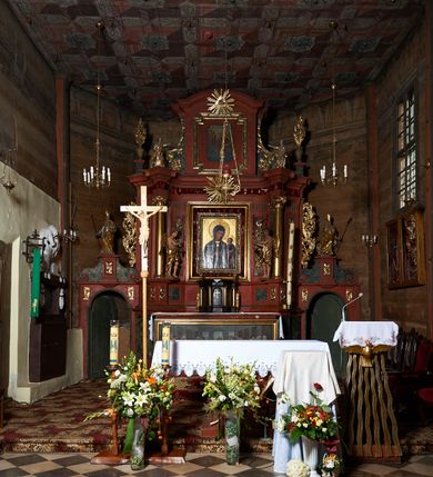 Zdjęcie nr 3:  Nastawa ołtarzowa architektoniczna, jednoosiowa, jednokondygnacyjna, na cokole, ze zwieńczeniem, z bramkami po bokach. Mensa drewniana z portatylem z marmuru dębnickiego, osadzona na murowanej, prostopadłościennej skrzyni. Antepedium w kształcie leżącego prostokąta, w profilowanej ramie z współczesnym obrazem Ostatnia Wieczerza, wzorowanym na fresku Leonarda da Vinci. Retabulum ujęte parą wklęsłych pilastrów oraz parą kolumn o kapitelach jońskich i gładkich trzonach, umieszczonych na ustawionych ukośnie cokołach. Podpory dźwigają pełne belkowanie o wklęsło-wypukłej linii, występujące ukośnie w osi kolumn. W polu środkowym profilowana rama w kształcie stojącego prostokąta. Zwieńczenie w kształcie stojącego prostokąta zamkniętego łukiem odcinkowym nadwieszonym i ujętego okazałymi spływami wolutowymi. W polu rama obrazu w formie stojącego prostokąta o wciętych narożach. Bramki z drzwiczkami, z przejściami w kształcie stojącego prostokąta zamkniętego łukiem półkolistym nadwieszonym, zwieńczone gzymsem, na którym w osi bramek cokoły ujęte spływami wolutowymi. Cokoły dekorowane płycinami z akantem w formie karbowanej wstęgi. Kolumny z dwóch stron flankują płyciny z festonami zwieńczone pasem z dekoracją geometryczną. W polu środkowym nastawy obraz Matka Boska z Dzieciątkiem otoczony wotami, w ramie dekorowanej ornamentem roślinnym, flankowany stojącymi przy kolumnach figurami aniołów. We fryzie wota, ponad polem środkowym gloria promienista ze splecionymi literami imienia MARIA. Nastawa ujęta uszami utworzonymi z motywów suchego akantu. Na belkowaniu dwie figury zwrócone do wnętrza ołtarza oraz cokoły z wazonami z płaskorzeźbionymi bukietami kwiatów. W zwieńczeniu rama dekorowana ornamentem roślinnym, w niej obraz Koronacja Marii , wyżej Oko Opatrzności w glorii. Spływy zdobione karbowaną wstęgą, akantem i kwiatami. Bramki w przyłuczach dekorowane akantem, cokół z płyciną i akantem, na nim figury św. Wojciecha i św. Stanisława. Nastawa polichromowana na czerwono, płyciny i detale architektoniczne marmoryzowane w odcieniach zieleni, ornamenty złocone.