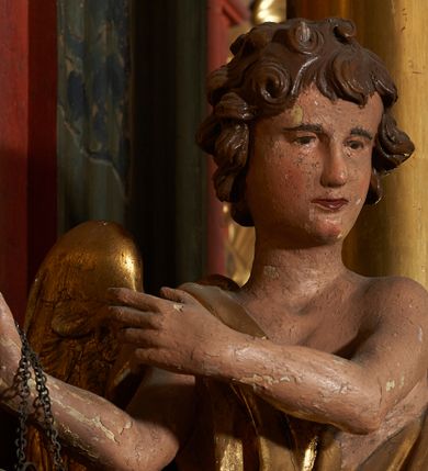 Zdjęcie nr 9: Rzeźby pełnoplastyczne, z tyłu drążone. Figury całopostaciowe, zwrócone antytetycznie, stojące w kontrapoście, z pochylonymi głowami, z obiema rękoma skierowanymi ku środkowi ołtarza, przytrzymują latarnie. Twarze owalne, o indywidualnych rysach, delikatnie zarysowanych oczach i ustach oraz płaskim nosie. Włosy w postaci krótkich, gęstych loków. Ubrane w przewiązaną diagonalnie przez tors, obficie drapowaną szatę, opadającą na tył postaci. Skrzydła złożone. Polichromia naturalistyczna w partiach ciała, szaty i skrzydła złocone.