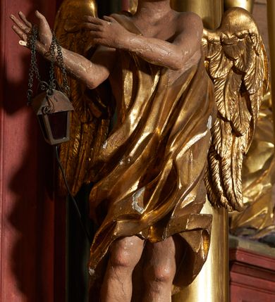 Zdjęcie nr 7: Rzeźby pełnoplastyczne, z tyłu drążone. Figury całopostaciowe, zwrócone antytetycznie, stojące w kontrapoście, z pochylonymi głowami, z obiema rękoma skierowanymi ku środkowi ołtarza, przytrzymują latarnie. Twarze owalne, o indywidualnych rysach, delikatnie zarysowanych oczach i ustach oraz płaskim nosie. Włosy w postaci krótkich, gęstych loków. Ubrane w przewiązaną diagonalnie przez tors, obficie drapowaną szatę, opadającą na tył postaci. Skrzydła złożone. Polichromia naturalistyczna w partiach ciała, szaty i skrzydła złocone.