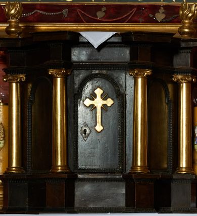Zdjęcie nr 1: Tabernakulum na rzucie połowy ośmioboku, opięte w narożach czterema kolumnami na cokołach, dźwigającymi pełne belkowanie i gzyms, występujące w osi podpór. Ścianki dekorowane płycinami w kształcie stojącego prostokąta zamkniętego łukiem półkolistym nadwieszonym. Drzwiczki o tym samym kształcie, osadzone na zawiasach, dekorowane aplikowanym krzyżem o zakończeniach ramion w kształcie rombu. Na gzymsie, w osi kolumn dwa wazony płomieniste. Całość w naturalnym odcieniu drewna, kolumny i detale złocone.