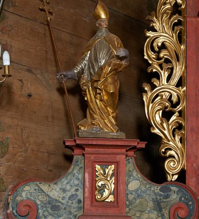 Zdjęcie nr 2: Rzeźba pełnoplastyczna, z tyłu drążona. Figura całopostaciowa, zwrócona en trois quarts w lewo, z głową skierowana w prawo, stojąca w kontrapoście i ekspresyjnym zwrocie ciała. Prawą ręką, wysuniętą w bok, podtrzymuje krzyż arcybiskupi, lewą ugiętą w łokciu trzyma połę kapy. Twarz szczupła, pociągła, o głęboko osadzonych oczach, dużym nosie i rozchylonych ustach, okolona długą, siwą brodą opadająca na pierś. Włosy krótkie, siwe, opadają na tył głowy. Święty ubrany w złoconą albę, srebrzoną komżę obficie drapowaną i skręconą wraz z postacią oraz ekspresyjnie rozwianą kapę, spiętą na piersi oraz zawiniętą na lewej nodze.