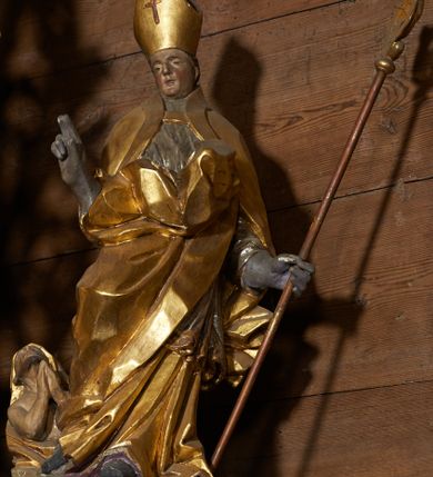 Zdjęcie nr 1: Rzeźba pełnoplastyczna, z tyłu drążona. Figura całopostaciowa, zwrócona en face do widza, stojąca w silnym kontrapoście, przechylona do tyłu. Prawą rękę wyciąga w geście błogosławieństwa, lewą odchyloną w bok, przytrzymuje pastorał. Twarz szczupła o wyraźnych rysach, głęboko osadzonych oczach, dużym nosie i niewielkich ustach. Na głowie infuła. Święty ubrany w złoconą albę, srebrzoną komżę, obficie drapowane, odpowiadające skrętowi sylwetki, oraz złoconą kapę spiętą na piersi, z ekspresyjnie rozwianym końcem przy prawym boku. Na dużych dłoniach rękawiczki. Przy prawej nodze świętego Piotrowin, ukazany w półpostaci, patrzący w górę, z rękami złożonymi w geście modlitwy.