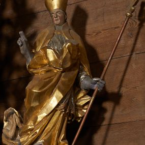 Zdjęcie nr 1: Rzeźba pełnoplastyczna, z tyłu drążona. Figura całopostaciowa, zwrócona en face do widza, stojąca w silnym kontrapoście, przechylona do tyłu. Prawą rękę wyciąga w geście błogosławieństwa, lewą odchyloną w bok, przytrzymuje pastorał. Twarz szczupła o wyraźnych rysach, głęboko osadzonych oczach, dużym nosie i niewielkich ustach. Na głowie infuła. Święty ubrany w złoconą albę, srebrzoną komżę, obficie drapowane, odpowiadające skrętowi sylwetki, oraz złoconą kapę spiętą na piersi, z ekspresyjnie rozwianym końcem przy prawym boku. Na dużych dłoniach rękawiczki. Przy prawej nodze świętego Piotrowin, ukazany w półpostaci, patrzący w górę, z rękami złożonymi w geście modlitwy.