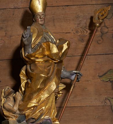 Zdjęcie nr 3: Rzeźba pełnoplastyczna, z tyłu drążona. Figura całopostaciowa, zwrócona en face do widza, stojąca w silnym kontrapoście, przechylona do tyłu. Prawą rękę wyciąga w geście błogosławieństwa, lewą odchyloną w bok, przytrzymuje pastorał. Twarz szczupła o wyraźnych rysach, głęboko osadzonych oczach, dużym nosie i niewielkich ustach. Na głowie infuła. Święty ubrany w złoconą albę, srebrzoną komżę, obficie drapowane, odpowiadające skrętowi sylwetki, oraz złoconą kapę spiętą na piersi, z ekspresyjnie rozwianym końcem przy prawym boku. Na dużych dłoniach rękawiczki. Przy prawej nodze świętego Piotrowin, ukazany w półpostaci, patrzący w górę, z rękami złożonymi w geście modlitwy.