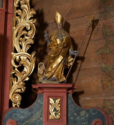 Zdjęcie nr 2: Rzeźba pełnoplastyczna, z tyłu drążona. Figura całopostaciowa, zwrócona en face do widza, stojąca w silnym kontrapoście, przechylona do tyłu. Prawą rękę wyciąga w geście błogosławieństwa, lewą odchyloną w bok, przytrzymuje pastorał. Twarz szczupła o wyraźnych rysach, głęboko osadzonych oczach, dużym nosie i niewielkich ustach. Na głowie infuła. Święty ubrany w złoconą albę, srebrzoną komżę, obficie drapowane, odpowiadające skrętowi sylwetki, oraz złoconą kapę spiętą na piersi, z ekspresyjnie rozwianym końcem przy prawym boku. Na dużych dłoniach rękawiczki. Przy prawej nodze świętego Piotrowin, ukazany w półpostaci, patrzący w górę, z rękami złożonymi w geście modlitwy.