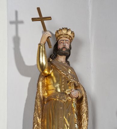 Zdjęcie nr 5: Pełnoplastyczna, drewniana figura św. Stefana, ustawiona na cokole na konsoli.  Święty został przedstawiony w pozycji stojącej, z lewą nogą wysuniętą nieco do przodu, ubrany w zapinany na guzki płaszcz i wysokie buty. Na to ma narzucony obfity płaszcz z futrzanym podbiciem spięty na prawym barku. Ma prosty nos i ciemne oczy. W prawej, uniesionej  ręce trzyma przed sobą krzyż, lewą opiera na rękojeści zatkniętego za pas miecza. Twarz pociągła, o wyrazistych rysach, okolona brodą. Spod zamkniętej korony spływają długie włosy opadające na oba ramiona i zwijające się w loki. Partie twarzy i włosów polichromowane naturalistycznie, szaty złocone.