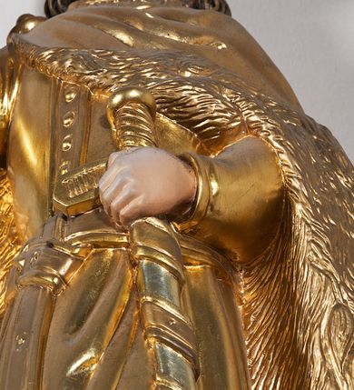 Zdjęcie nr 8: Pełnoplastyczna, drewniana figura św. Stefana, ustawiona na cokole na konsoli.  Święty został przedstawiony w pozycji stojącej, z lewą nogą wysuniętą nieco do przodu, ubrany w zapinany na guzki płaszcz i wysokie buty. Na to ma narzucony obfity płaszcz z futrzanym podbiciem spięty na prawym barku. Ma prosty nos i ciemne oczy. W prawej, uniesionej  ręce trzyma przed sobą krzyż, lewą opiera na rękojeści zatkniętego za pas miecza. Twarz pociągła, o wyrazistych rysach, okolona brodą. Spod zamkniętej korony spływają długie włosy opadające na oba ramiona i zwijające się w loki. Partie twarzy i włosów polichromowane naturalistycznie, szaty złocone.