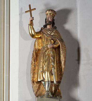 Zdjęcie nr 1: Pełnoplastyczna, drewniana figura św. Stefana, ustawiona na cokole na konsoli.  Święty został przedstawiony w pozycji stojącej, z lewą nogą wysuniętą nieco do przodu, ubrany w zapinany na guzki płaszcz i wysokie buty. Na to ma narzucony obfity płaszcz z futrzanym podbiciem spięty na prawym barku. Ma prosty nos i ciemne oczy. W prawej, uniesionej  ręce trzyma przed sobą krzyż, lewą opiera na rękojeści zatkniętego za pas miecza. Twarz pociągła, o wyrazistych rysach, okolona brodą. Spod zamkniętej korony spływają długie włosy opadające na oba ramiona i zwijające się w loki. Partie twarzy i włosów polichromowane naturalistycznie, szaty złocone.