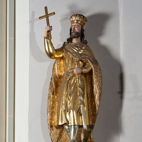 Zdjęcie nr 1: Pełnoplastyczna, drewniana figura św. Stefana, ustawiona na cokole na konsoli.  Święty został przedstawiony w pozycji stojącej, z lewą nogą wysuniętą nieco do przodu, ubrany w zapinany na guzki płaszcz i wysokie buty. Na to ma narzucony obfity płaszcz z futrzanym podbiciem spięty na prawym barku. Ma prosty nos i ciemne oczy. W prawej, uniesionej  ręce trzyma przed sobą krzyż, lewą opiera na rękojeści zatkniętego za pas miecza. Twarz pociągła, o wyrazistych rysach, okolona brodą. Spod zamkniętej korony spływają długie włosy opadające na oba ramiona i zwijające się w loki. Partie twarzy i włosów polichromowane naturalistycznie, szaty złocone.