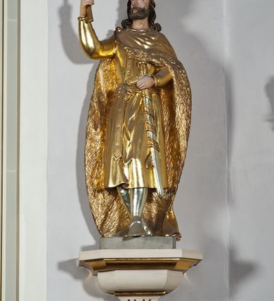 Zdjęcie nr 3: Pełnoplastyczna, drewniana figura św. Stefana, ustawiona na cokole na konsoli.  Święty został przedstawiony w pozycji stojącej, z lewą nogą wysuniętą nieco do przodu, ubrany w zapinany na guzki płaszcz i wysokie buty. Na to ma narzucony obfity płaszcz z futrzanym podbiciem spięty na prawym barku. Ma prosty nos i ciemne oczy. W prawej, uniesionej  ręce trzyma przed sobą krzyż, lewą opiera na rękojeści zatkniętego za pas miecza. Twarz pociągła, o wyrazistych rysach, okolona brodą. Spod zamkniętej korony spływają długie włosy opadające na oba ramiona i zwijające się w loki. Partie twarzy i włosów polichromowane naturalistycznie, szaty złocone.