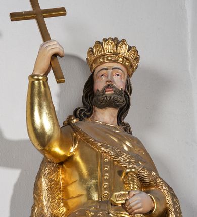 Zdjęcie nr 6: Pełnoplastyczna, drewniana figura św. Stefana, ustawiona na cokole na konsoli.  Święty został przedstawiony w pozycji stojącej, z lewą nogą wysuniętą nieco do przodu, ubrany w zapinany na guzki płaszcz i wysokie buty. Na to ma narzucony obfity płaszcz z futrzanym podbiciem spięty na prawym barku. Ma prosty nos i ciemne oczy. W prawej, uniesionej  ręce trzyma przed sobą krzyż, lewą opiera na rękojeści zatkniętego za pas miecza. Twarz pociągła, o wyrazistych rysach, okolona brodą. Spod zamkniętej korony spływają długie włosy opadające na oba ramiona i zwijające się w loki. Partie twarzy i włosów polichromowane naturalistycznie, szaty złocone.