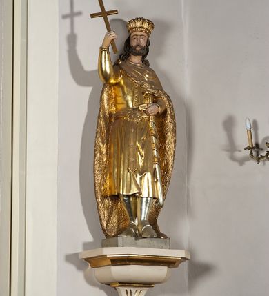 Zdjęcie nr 2: Pełnoplastyczna, drewniana figura św. Stefana, ustawiona na cokole na konsoli.  Święty został przedstawiony w pozycji stojącej, z lewą nogą wysuniętą nieco do przodu, ubrany w zapinany na guzki płaszcz i wysokie buty. Na to ma narzucony obfity płaszcz z futrzanym podbiciem spięty na prawym barku. Ma prosty nos i ciemne oczy. W prawej, uniesionej  ręce trzyma przed sobą krzyż, lewą opiera na rękojeści zatkniętego za pas miecza. Twarz pociągła, o wyrazistych rysach, okolona brodą. Spod zamkniętej korony spływają długie włosy opadające na oba ramiona i zwijające się w loki. Partie twarzy i włosów polichromowane naturalistycznie, szaty złocone.