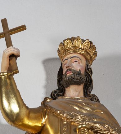 Zdjęcie nr 7: Pełnoplastyczna, drewniana figura św. Stefana, ustawiona na cokole na konsoli.  Święty został przedstawiony w pozycji stojącej, z lewą nogą wysuniętą nieco do przodu, ubrany w zapinany na guzki płaszcz i wysokie buty. Na to ma narzucony obfity płaszcz z futrzanym podbiciem spięty na prawym barku. Ma prosty nos i ciemne oczy. W prawej, uniesionej  ręce trzyma przed sobą krzyż, lewą opiera na rękojeści zatkniętego za pas miecza. Twarz pociągła, o wyrazistych rysach, okolona brodą. Spod zamkniętej korony spływają długie włosy opadające na oba ramiona i zwijające się w loki. Partie twarzy i włosów polichromowane naturalistycznie, szaty złocone.