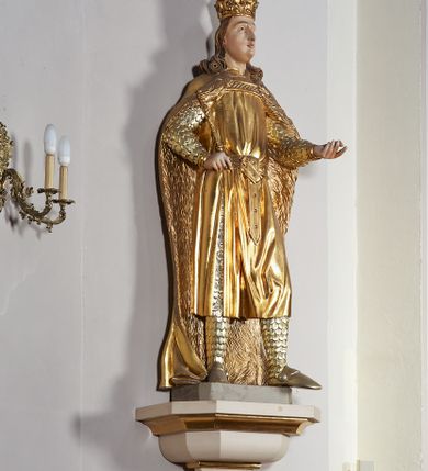 Zdjęcie nr 7: Pełnoplastyczna, drewniana figura św. Ludwika, ustawiona na konsoli. Święty został przedstawiony w pozycji stojącej, z lewą nogą wysuniętą nieco do przodu, ubrany w zbroję łuskową, na którą narzucono mu przewiązaną pasem tunikę i obfity płaszcz z futrzanym podbiciem spięty pod szyją. Prawą, ugiętą rękę opiera na biodrze, lewą wyciąga przed siebie. Twarz pociągła, okolona długimi włosami opadającymi na oba ramiona i zwijającymi się w loki. Ma niewielkie, pełne usta, prosty nos i ciemne oczy; na głowie korona zamknięta. Partie twarzy i włosów polichromowane naturalistycznie, szaty złocone. 