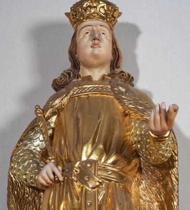 Zdjęcie nr 2: Pełnoplastyczna, drewniana figura św. Ludwika, ustawiona na konsoli. Święty został przedstawiony w pozycji stojącej, z lewą nogą wysuniętą nieco do przodu, ubrany w zbroję łuskową, na którą narzucono mu przewiązaną pasem tunikę i obfity płaszcz z futrzanym podbiciem spięty pod szyją. Prawą, ugiętą rękę opiera na biodrze, lewą wyciąga przed siebie. Twarz pociągła, okolona długimi włosami opadającymi na oba ramiona i zwijającymi się w loki. Ma niewielkie, pełne usta, prosty nos i ciemne oczy; na głowie korona zamknięta. Partie twarzy i włosów polichromowane naturalistycznie, szaty złocone. 