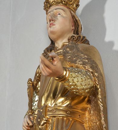 Zdjęcie nr 4: Pełnoplastyczna, drewniana figura św. Ludwika, ustawiona na konsoli. Święty został przedstawiony w pozycji stojącej, z lewą nogą wysuniętą nieco do przodu, ubrany w zbroję łuskową, na którą narzucono mu przewiązaną pasem tunikę i obfity płaszcz z futrzanym podbiciem spięty pod szyją. Prawą, ugiętą rękę opiera na biodrze, lewą wyciąga przed siebie. Twarz pociągła, okolona długimi włosami opadającymi na oba ramiona i zwijającymi się w loki. Ma niewielkie, pełne usta, prosty nos i ciemne oczy; na głowie korona zamknięta. Partie twarzy i włosów polichromowane naturalistycznie, szaty złocone. 