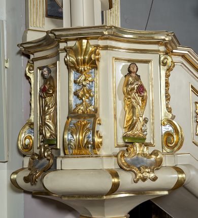 Zdjęcie nr 6: Ambona przyścienna, wisząca, dostępna schodami z podestem. Ich balustrada jest pełna dekorowana płycinami wypełnionymi plakietami z ornamentacją roślinną. Kosz na profilowanym cokole z półwałkiem, na planie prostokąta o wklęsło-wypukłych ściankach wydzielonych spływami z wolutami, dekorowanymi kampanulami i liśćmi akantu. Na wklęsłych ściankach płyciny w kształcie stojącego prostokąta, wypełnione płaskorzeźbionymi, całopostaciowymi przedstawieniami św. Łukasza z lewej i św. Jana Ewangelisty z prawej strony, pod ich stopami kartusze ujęte dekoracją rocaille’ową.  Parapet w postaci profilowanego gzymsu wyłamanego nad spływami. Spód kosza w kształcie wklęsło-wypukłego, odwróconego stożka zakończonego szyszką. Zaplecek w formie wydłużonego prostokąta ujętego kanelowanymi pilastrami. W polu obraz w kształcie stojącego prostokąta z wizerunkiem Chrystusa Dobrego Pasterza. Pilastry podtrzymują profilowane belkowanie wyłamane na ich osiach, na których znajdują się fragmenty przyczółka skierowane na zewnątrz. Między nimi, na ścianie rozbudowane przedstawienie gołębicy Ducha Świętego na tle glorii promienistej  i obłoków.  Struktura w odcieniu kremowym, dekoracja i profile złocone i srebrzone. 