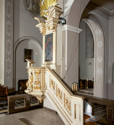 Zdjęcie nr 3: Ambona przyścienna, wisząca, dostępna schodami z podestem. Ich balustrada jest pełna dekorowana płycinami wypełnionymi plakietami z ornamentacją roślinną. Kosz na profilowanym cokole z półwałkiem, na planie prostokąta o wklęsło-wypukłych ściankach wydzielonych spływami z wolutami, dekorowanymi kampanulami i liśćmi akantu. Na wklęsłych ściankach płyciny w kształcie stojącego prostokąta, wypełnione płaskorzeźbionymi, całopostaciowymi przedstawieniami św. Łukasza z lewej i św. Jana Ewangelisty z prawej strony, pod ich stopami kartusze ujęte dekoracją rocaille’ową.  Parapet w postaci profilowanego gzymsu wyłamanego nad spływami. Spód kosza w kształcie wklęsło-wypukłego, odwróconego stożka zakończonego szyszką. Zaplecek w formie wydłużonego prostokąta ujętego kanelowanymi pilastrami. W polu obraz w kształcie stojącego prostokąta z wizerunkiem Chrystusa Dobrego Pasterza. Pilastry podtrzymują profilowane belkowanie wyłamane na ich osiach, na których znajdują się fragmenty przyczółka skierowane na zewnątrz. Między nimi, na ścianie rozbudowane przedstawienie gołębicy Ducha Świętego na tle glorii promienistej  i obłoków.  Struktura w odcieniu kremowym, dekoracja i profile złocone i srebrzone. 
