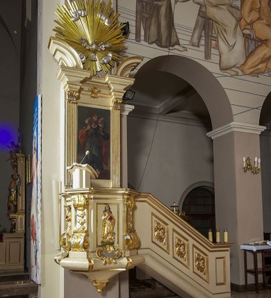 Zdjęcie nr 1: Ambona przyścienna, wisząca, dostępna schodami z podestem. Ich balustrada jest pełna dekorowana płycinami wypełnionymi plakietami z ornamentacją roślinną. Kosz na profilowanym cokole z półwałkiem, na planie prostokąta o wklęsło-wypukłych ściankach wydzielonych spływami z wolutami, dekorowanymi kampanulami i liśćmi akantu. Na wklęsłych ściankach płyciny w kształcie stojącego prostokąta, wypełnione płaskorzeźbionymi, całopostaciowymi przedstawieniami św. Łukasza z lewej i św. Jana Ewangelisty z prawej strony, pod ich stopami kartusze ujęte dekoracją rocaille’ową.  Parapet w postaci profilowanego gzymsu wyłamanego nad spływami. Spód kosza w kształcie wklęsło-wypukłego, odwróconego stożka zakończonego szyszką. Zaplecek w formie wydłużonego prostokąta ujętego kanelowanymi pilastrami. W polu obraz w kształcie stojącego prostokąta z wizerunkiem Chrystusa Dobrego Pasterza. Pilastry podtrzymują profilowane belkowanie wyłamane na ich osiach, na których znajdują się fragmenty przyczółka skierowane na zewnątrz. Między nimi, na ścianie rozbudowane przedstawienie gołębicy Ducha Świętego na tle glorii promienistej  i obłoków.  Struktura w odcieniu kremowym, dekoracja i profile złocone i srebrzone. 