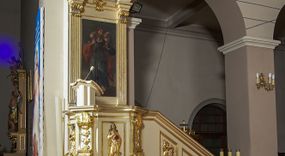 Zdjęcie nr 1: Ambona przyścienna, wisząca, dostępna schodami z podestem. Ich balustrada jest pełna dekorowana płycinami wypełnionymi plakietami z ornamentacją roślinną. Kosz na profilowanym cokole z półwałkiem, na planie prostokąta o wklęsło-wypukłych ściankach wydzielonych spływami z wolutami, dekorowanymi kampanulami i liśćmi akantu. Na wklęsłych ściankach płyciny w kształcie stojącego prostokąta, wypełnione płaskorzeźbionymi, całopostaciowymi przedstawieniami św. Łukasza z lewej i św. Jana Ewangelisty z prawej strony, pod ich stopami kartusze ujęte dekoracją rocaille’ową.  Parapet w postaci profilowanego gzymsu wyłamanego nad spływami. Spód kosza w kształcie wklęsło-wypukłego, odwróconego stożka zakończonego szyszką. Zaplecek w formie wydłużonego prostokąta ujętego kanelowanymi pilastrami. W polu obraz w kształcie stojącego prostokąta z wizerunkiem Chrystusa Dobrego Pasterza. Pilastry podtrzymują profilowane belkowanie wyłamane na ich osiach, na których znajdują się fragmenty przyczółka skierowane na zewnątrz. Między nimi, na ścianie rozbudowane przedstawienie gołębicy Ducha Świętego na tle glorii promienistej  i obłoków.  Struktura w odcieniu kremowym, dekoracja i profile złocone i srebrzone. 