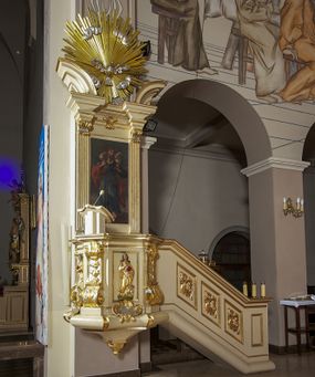 Zdjęcie nr 1: Ambona przyścienna, wisząca, dostępna schodami z podestem. Ich balustrada jest pełna dekorowana płycinami wypełnionymi plakietami z ornamentacją roślinną. Kosz na profilowanym cokole z półwałkiem, na planie prostokąta o wklęsło-wypukłych ściankach wydzielonych spływami z wolutami, dekorowanymi kampanulami i liśćmi akantu. Na wklęsłych ściankach płyciny w kształcie stojącego prostokąta, wypełnione płaskorzeźbionymi, całopostaciowymi przedstawieniami św. Łukasza z lewej i św. Jana Ewangelisty z prawej strony, pod ich stopami kartusze ujęte dekoracją rocaille’ową.  Parapet w postaci profilowanego gzymsu wyłamanego nad spływami. Spód kosza w kształcie wklęsło-wypukłego, odwróconego stożka zakończonego szyszką. Zaplecek w formie wydłużonego prostokąta ujętego kanelowanymi pilastrami. W polu obraz w kształcie stojącego prostokąta z wizerunkiem Chrystusa Dobrego Pasterza. Pilastry podtrzymują profilowane belkowanie wyłamane na ich osiach, na których znajdują się fragmenty przyczółka skierowane na zewnątrz. Między nimi, na ścianie rozbudowane przedstawienie gołębicy Ducha Świętego na tle glorii promienistej  i obłoków.  Struktura w odcieniu kremowym, dekoracja i profile złocone i srebrzone. 